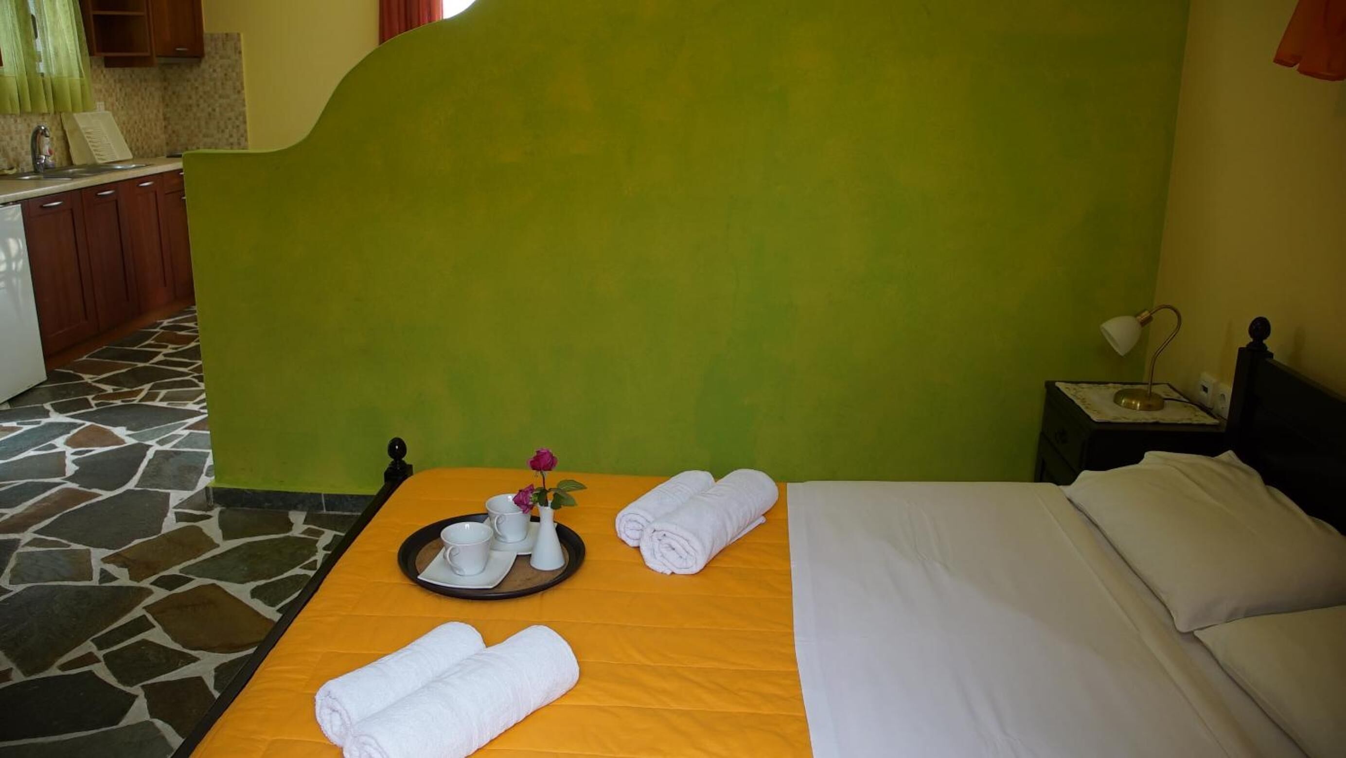 Una habitación de hotel con cama, una mesita de noche con lámpara, una pequeña mesa con un florero y un baño a la vista al fondo.