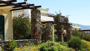 A building with stone pillars, a pergola, and a garden with yellow flowers and red blooms. - Ktima Nikola (Karystos)