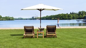 A lakeside setting with two lounge chairs and a table under a large umbrella, overlooking a calm lake and a distant cityscape.
