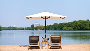 A beach setting with two wooden lounge chairs under a white umbrella, a small table with a bowl and a bottle, and a calm lake with a distant cityscape.
