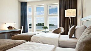 A hotel room with two beds, a desk, a lamp, and a view of the sea.