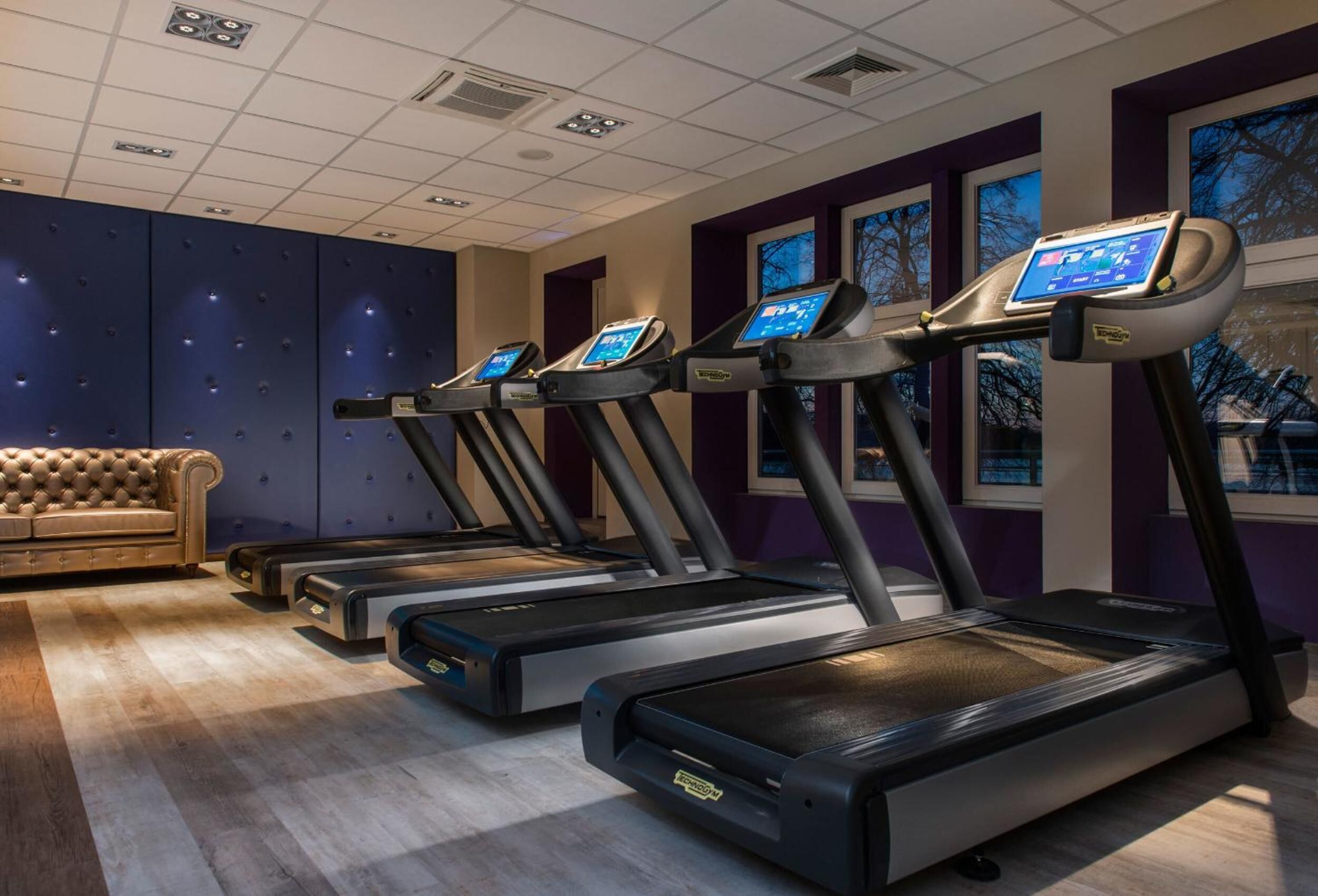 Ein Fitnessraum mit vier Laufbändern, einem bequemen Sofa und großen Fenstern.