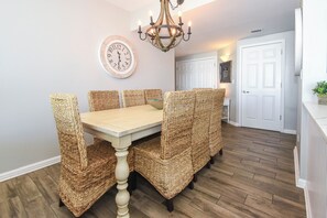 A dining area with a wooden table and wicker chairs, a wall clock, and a chandelier. - Remodeled, elegant beachfront condo w/private balcony! 🐚 (Destin)