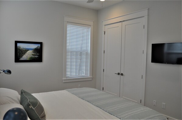 A bedroom with a bed, a window with blinds, a door, a television mounted on the wall, and a painting on the wall.