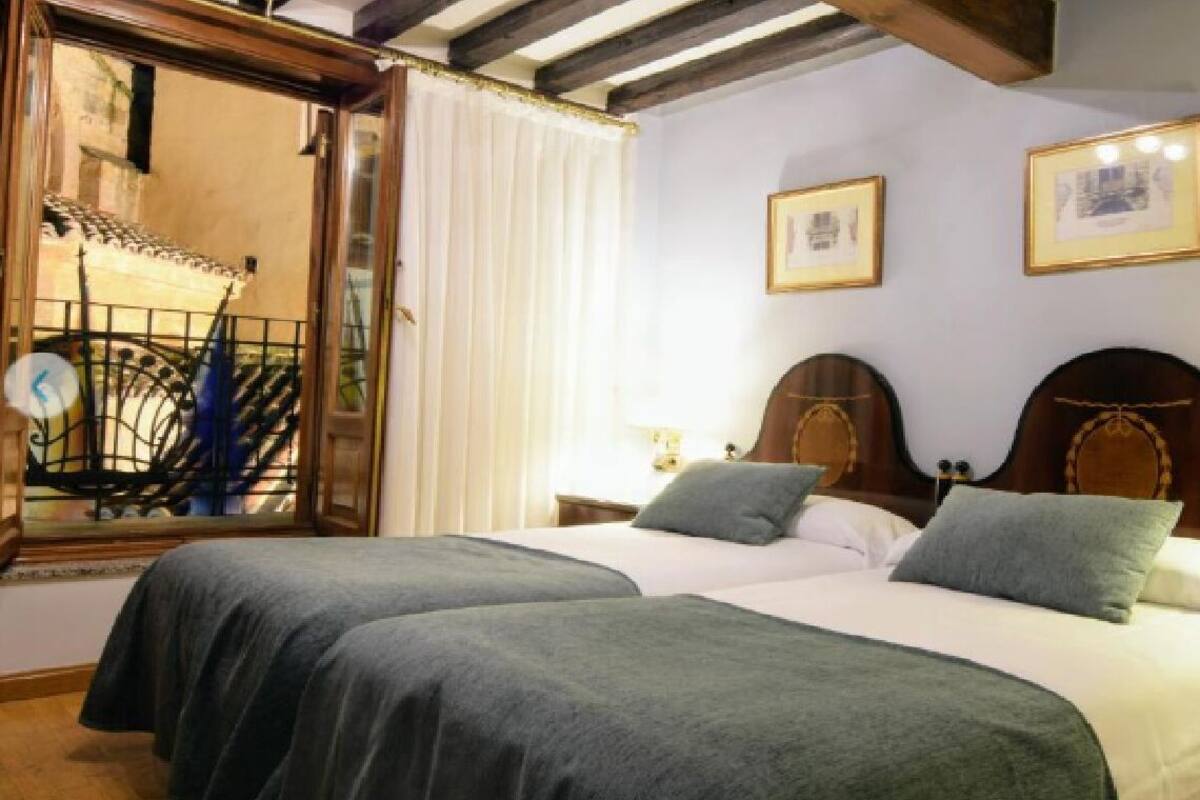 A room with two beds, wooden headboards, a window with a view, and framed pictures on the wall.