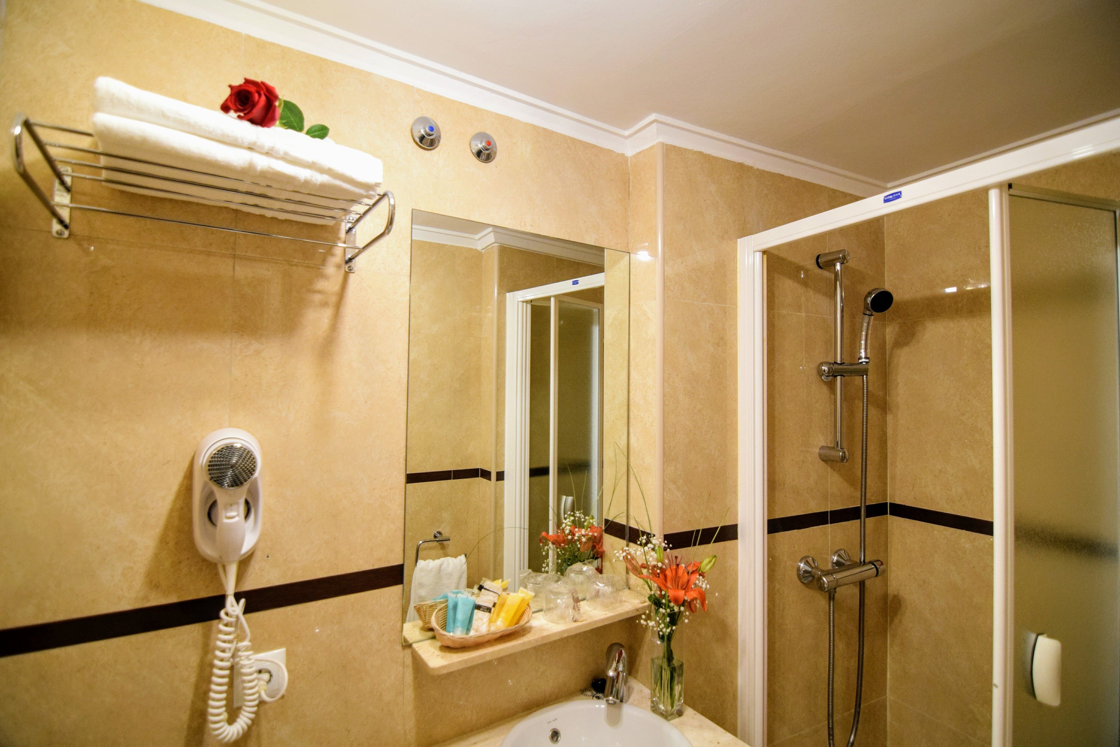 Un baño con lavamanos, espejo, toallero con toalla y una rosa, secador de pelo, ducha y estante con artículos de aseo y flores.