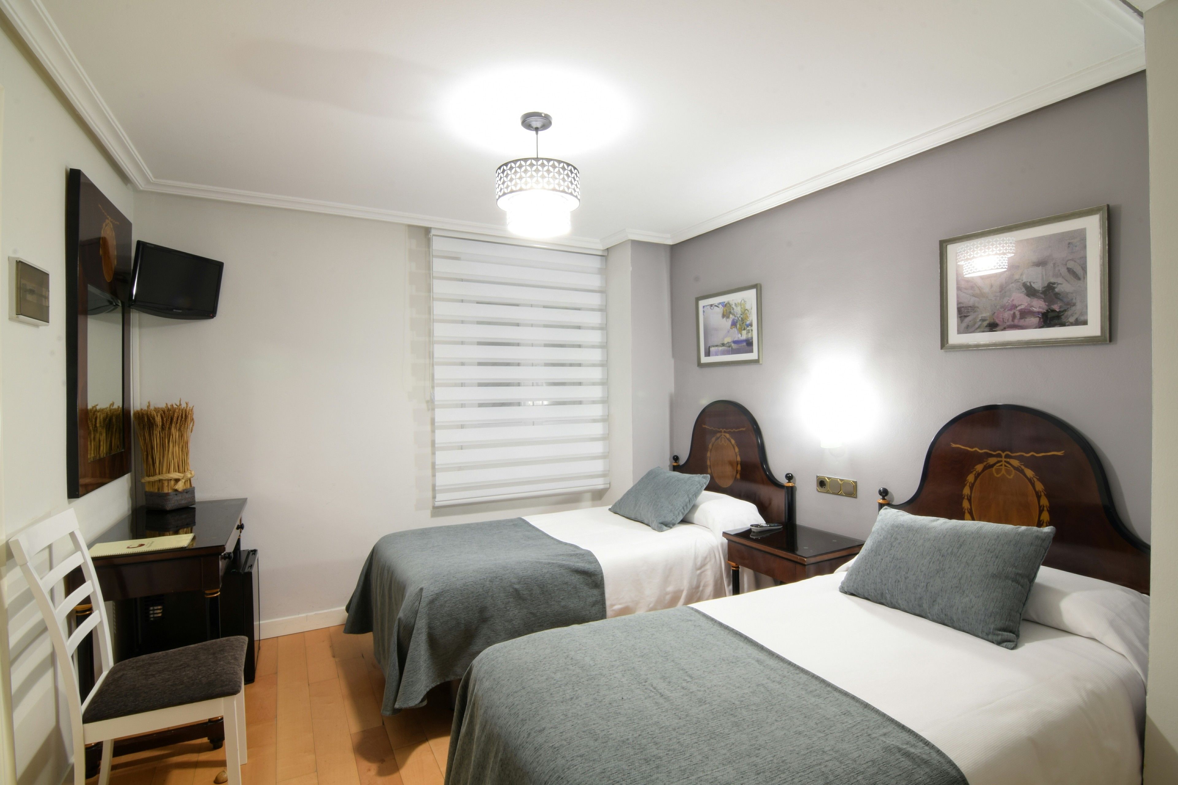 A bedroom with two beds, a desk, a chair, a TV, and a window with blinds.