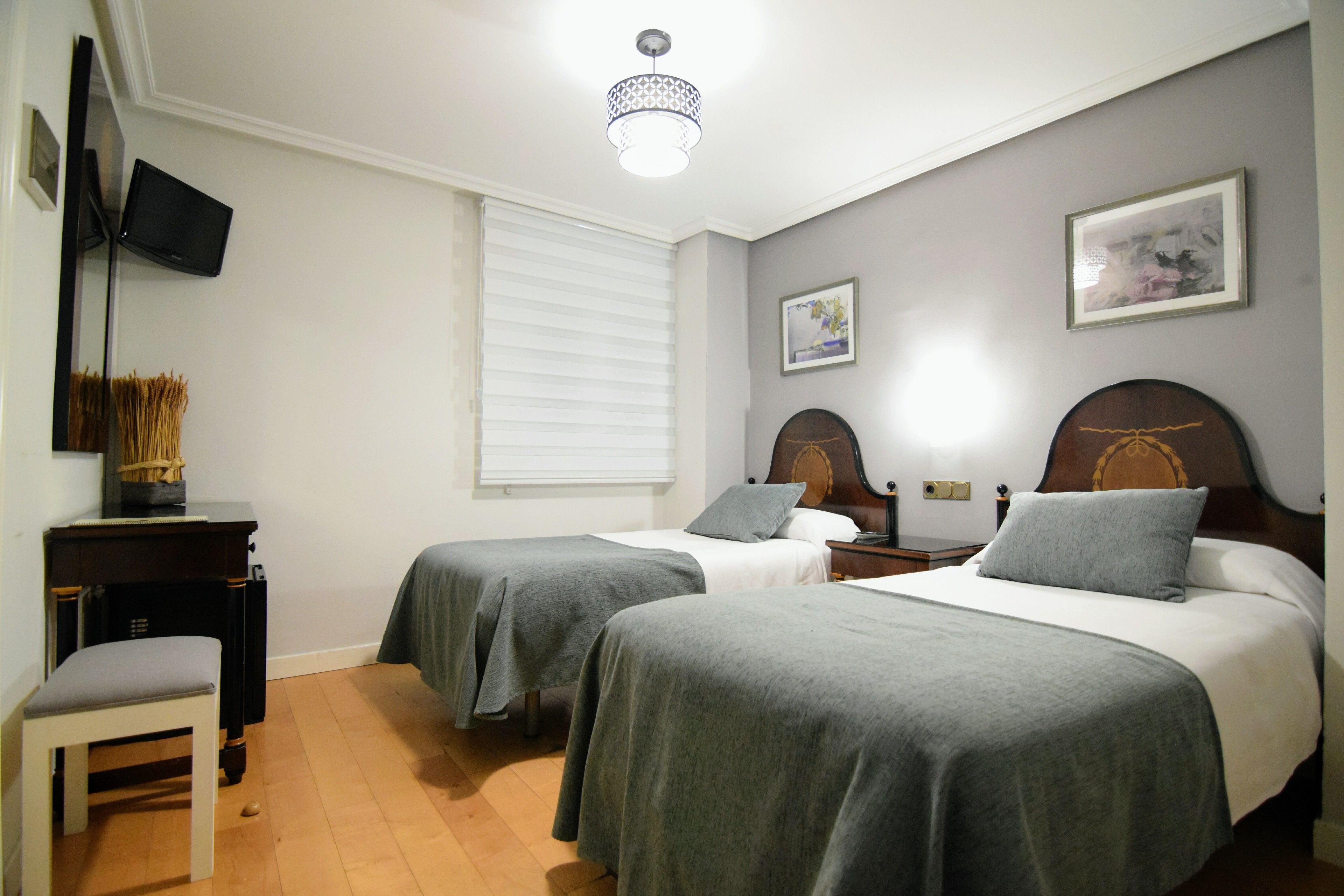 Habitación de hotel con dos camas individuales, un escritorio con silla, televisor de pantalla plana y lámpara en el techo.