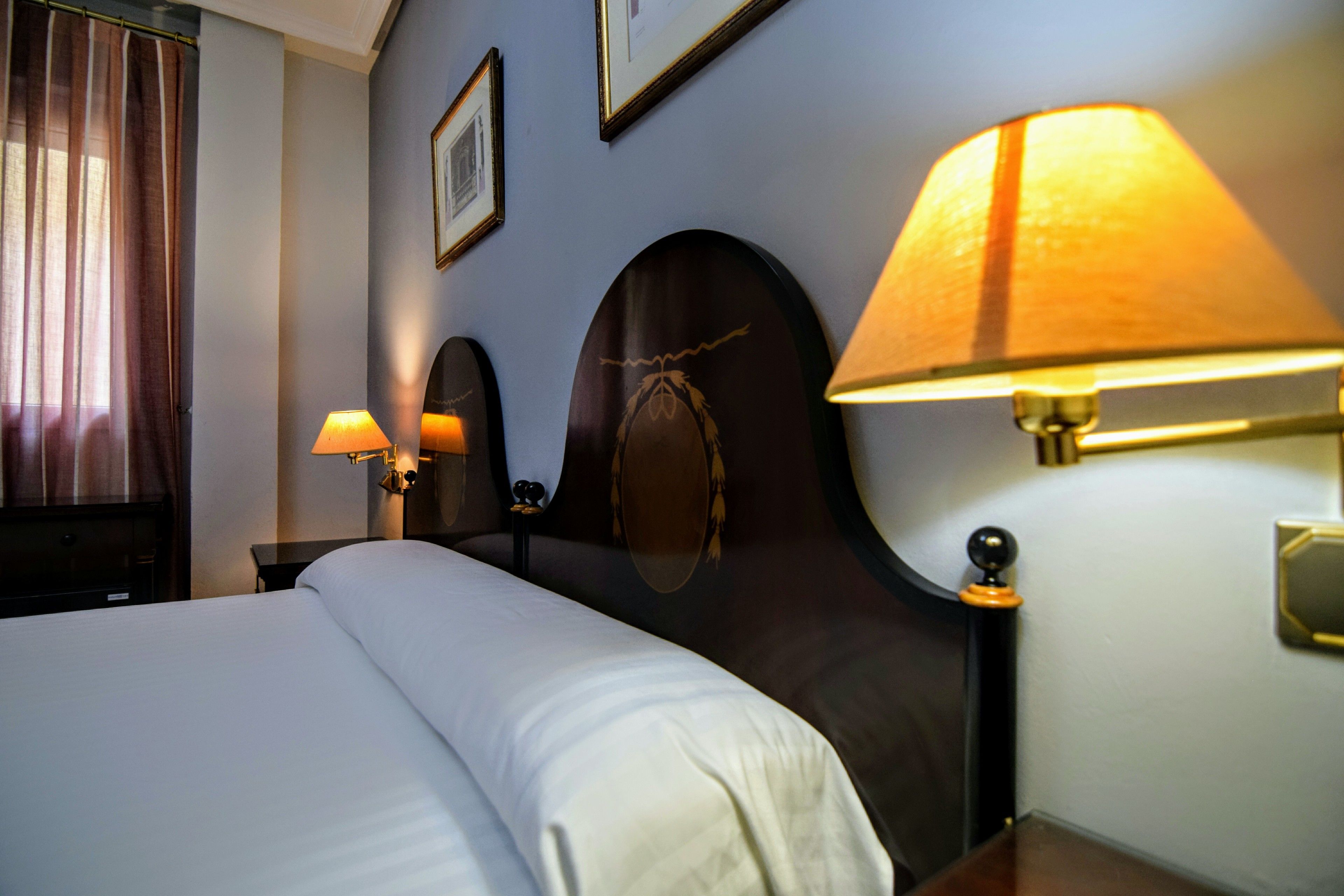 A hotel room with a large bed, two bedside lamps, a headboard with a mirror, and a window with curtains.