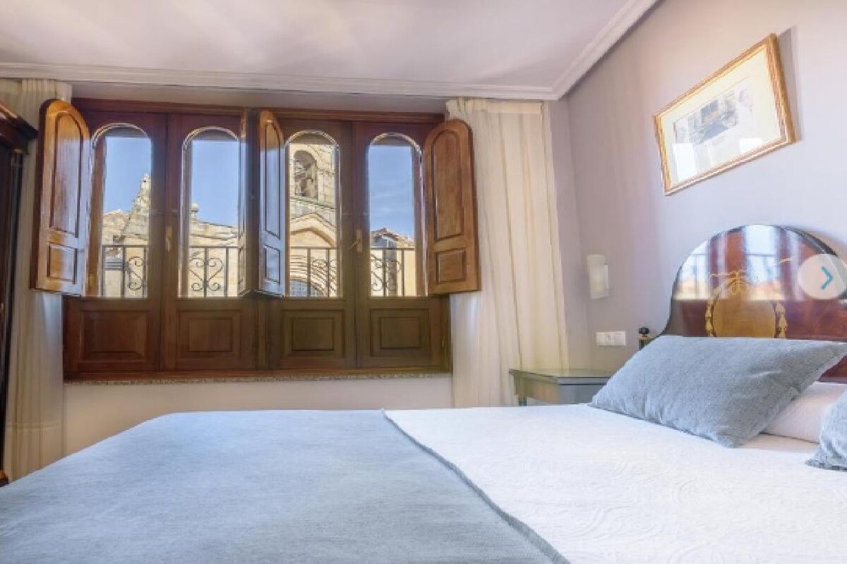 A bedroom with a bed, a nightstand, a clock, and a window with a view of a building.