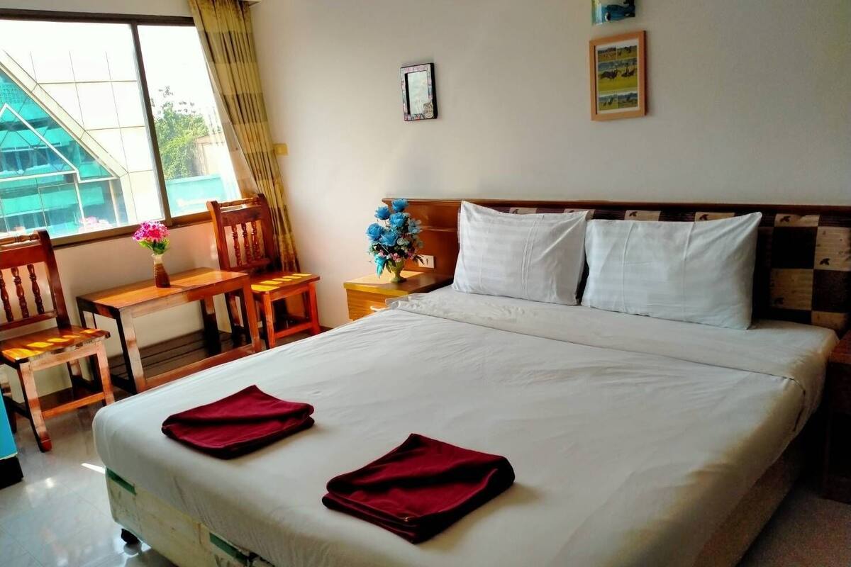 Une chambre d’hôtel avec un grand lit, un bureau en bois, deux chaises, une petite table ornée d’un vase de fleurs et une fenêtre avec des rideaux.