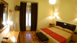 Une chambre d’hôtel avec un plancher en bois, un lit recouvert d’une couverture rouge, un banc, un bureau avec une lampe, un miroir, une télévision et une fenêtre avec des rideaux.