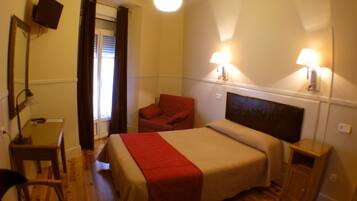 Habitación de hotel con cama, escritorio, silla, televisor y dos lámparas fijadas a la pared.