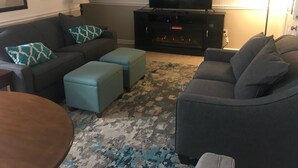 Una sala de estar con sofá gris, almohadones estampados en color verde azulado, un televisor de pantalla plana, chimenea y alfombra con diseño.