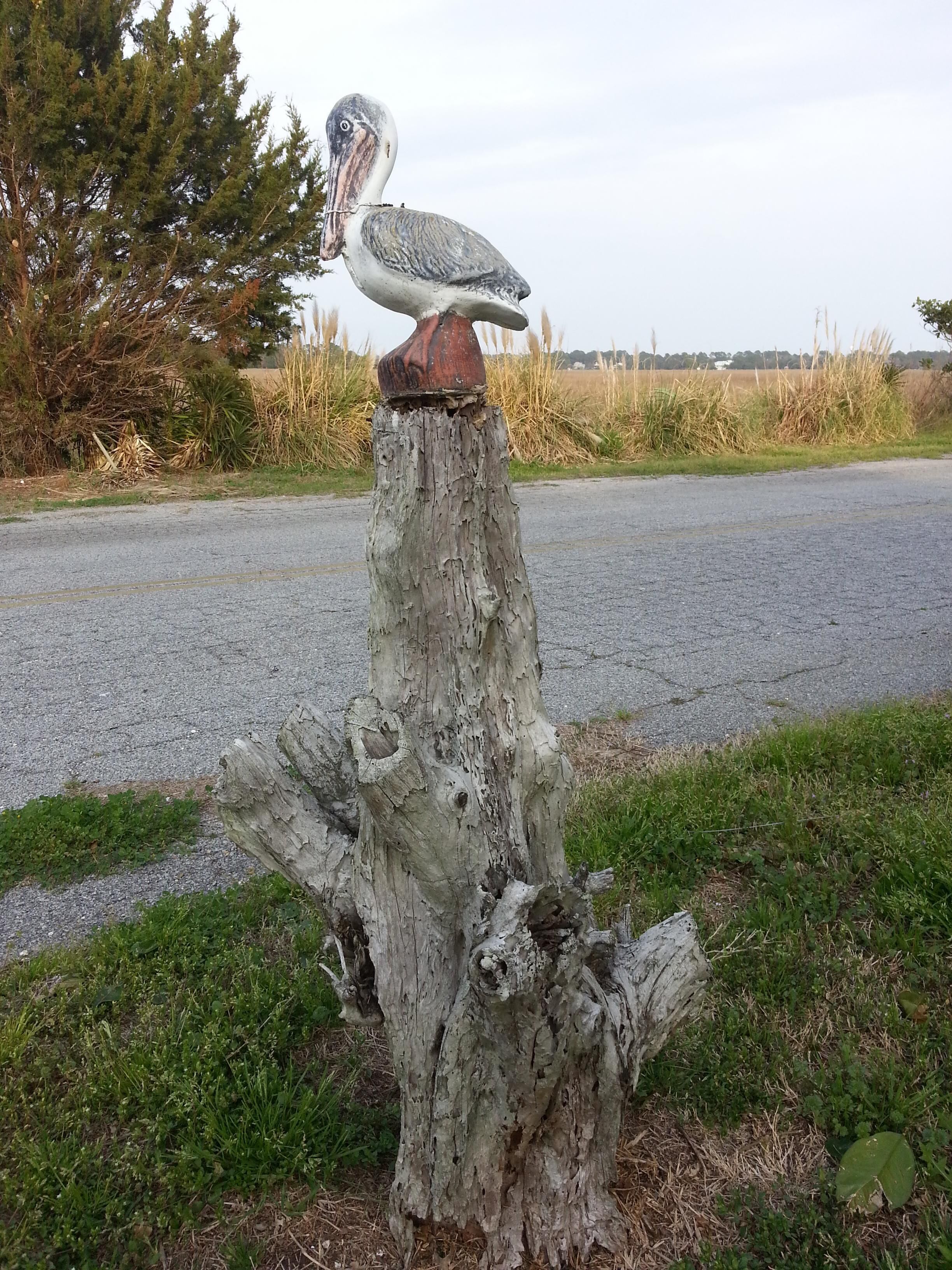 A wooden post with a bird sculpture on top, surrounded by grass and a road in the background.