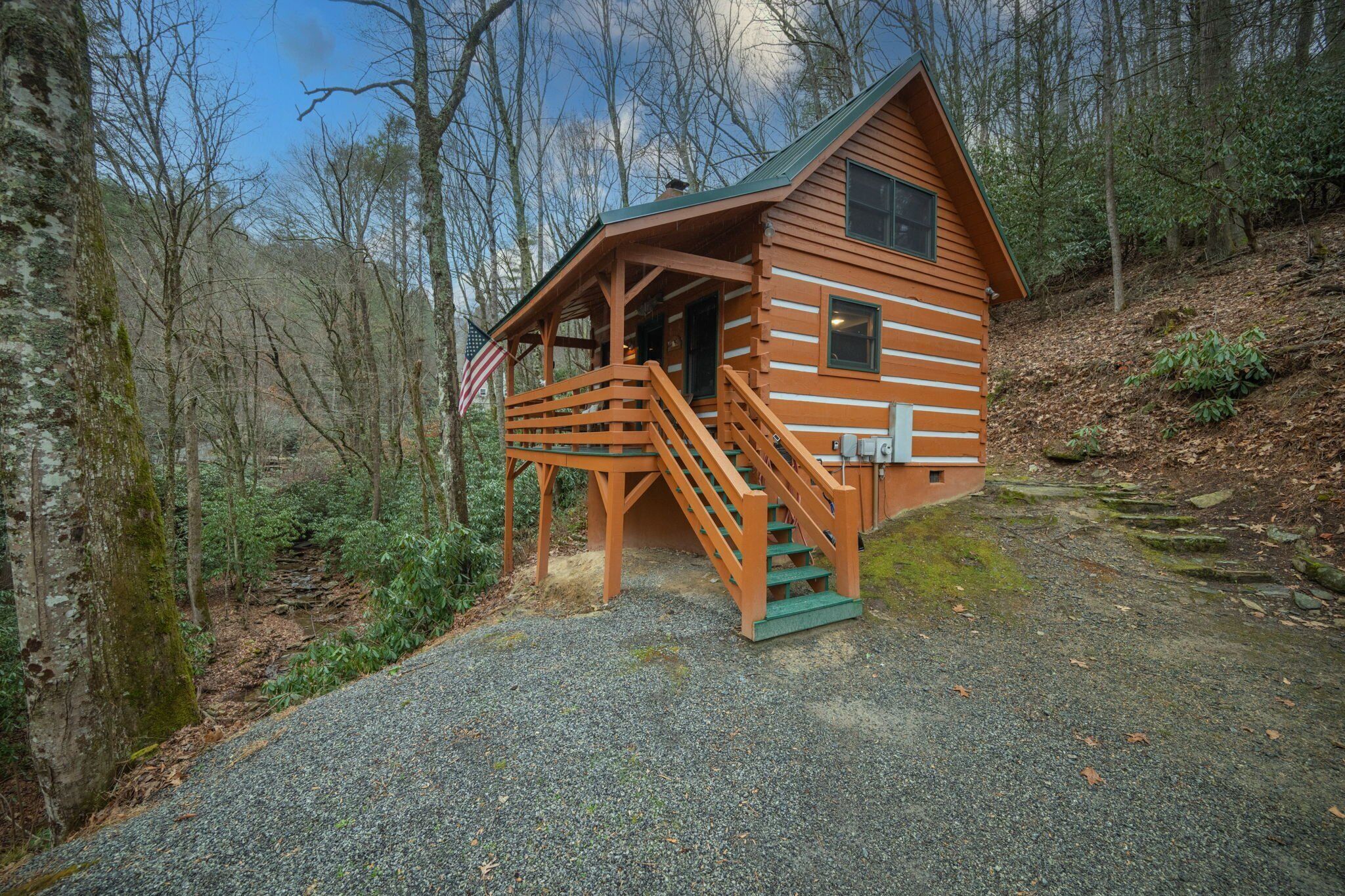A wooden cabin with a porch and stairs, surrounded by trees and a gravel path.