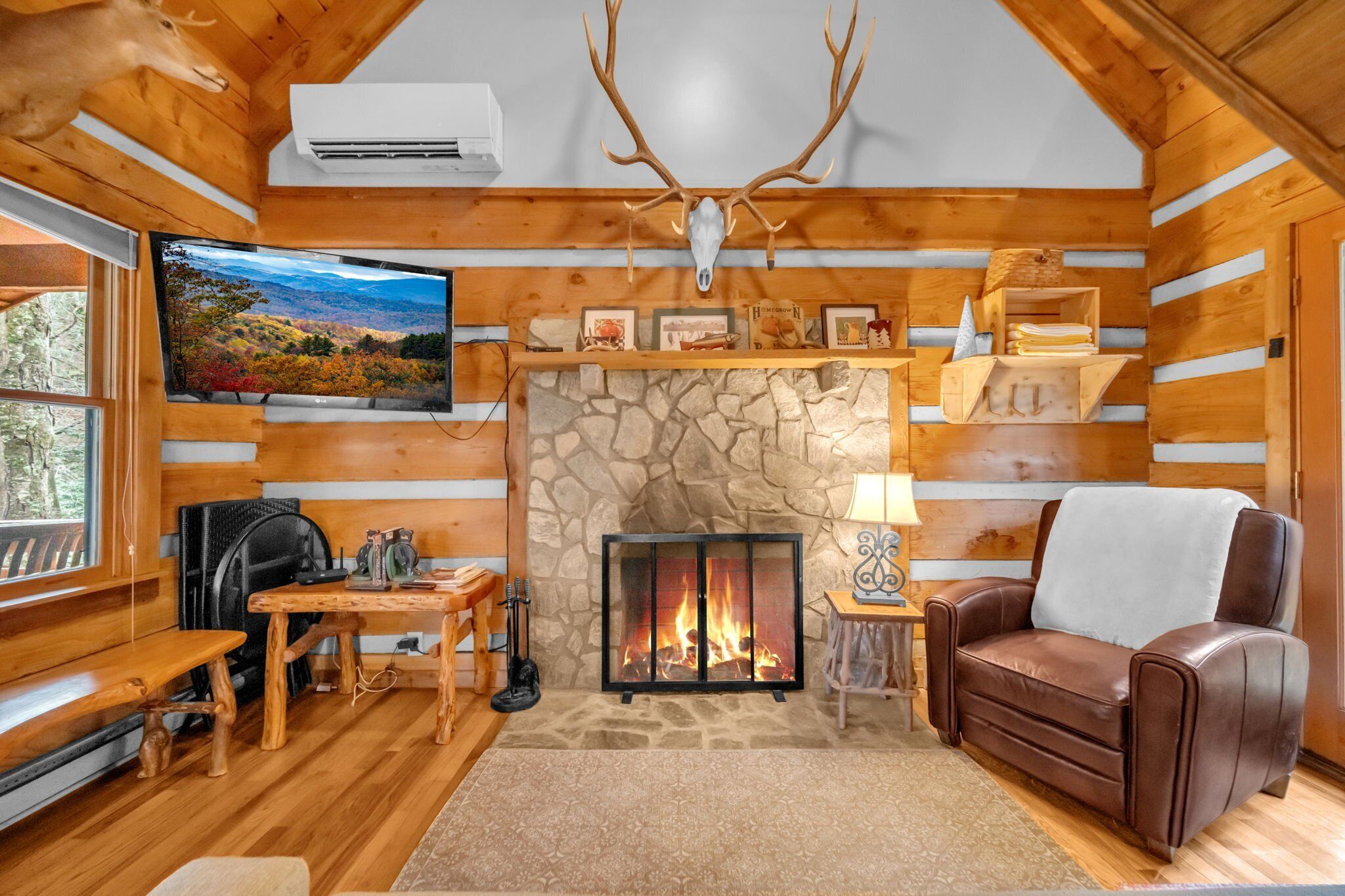 Ein gemütliches Blockhaus-Interieur mit Kamin aus Stein, einem aufgehängten Hirschgeweih, einem Fernseher und einem Ledersofa.