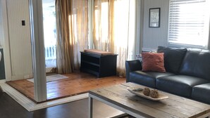 A living room with a dark wood floor, a white-framed coffee table, a black leather sofa with a red pillow, and a fish decoration on the wall. - The Buccaneer-Cottage, great for a couple or small family. (Ocracoke)