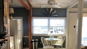 A kitchen with a refrigerator, a dining table with chairs, a window with blinds, and a ceiling fan. - The Buccaneer-Cottage, great for a couple or small family. (Ocracoke)