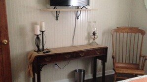 A bedroom with a bed, a wooden chair, a small table with candles, a television mounted on the wall, and a trash can. - The Buccaneer-Cottage, great for a couple or small family. (Ocracoke)