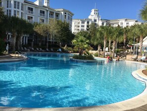 A large, clear swimming pool surrounded by palm trees and lounge chairs, with a multi-story building in the background. - Spectacular view, Big corner balcony, Lagoon pool, Baytowne & free shuttle (Miramar Beach)
