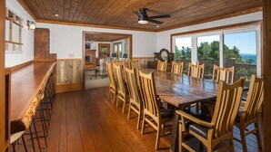 Un vaste espace dédié à la salle à manger, avec une longue table en bois et des chaises, un coin bar avec des tabourets, et de grandes fenêtres offrant une vue sur l’extérieur.