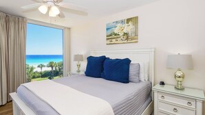 A bedroom with a bed, a nightstand, a lamp, a ceiling fan, a painting, and a view of the ocean.