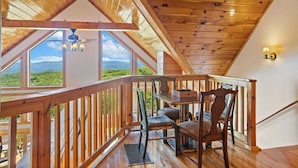 A wooden deck with a table and chairs, a view of green hills through large windows, and a sloped ceiling.