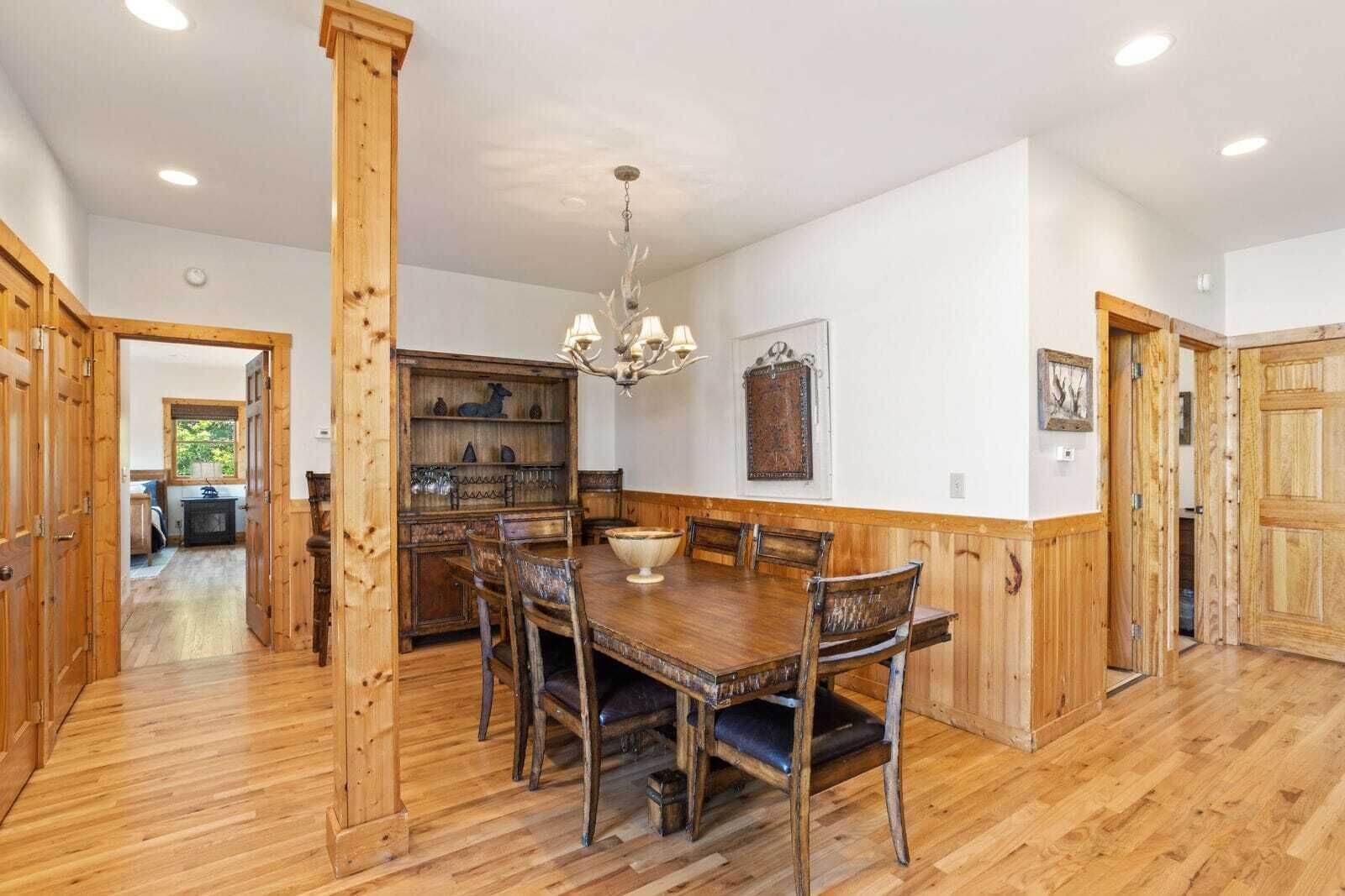 A dining area with a wooden table and chairs, a chandelier, and a wooden cabinet with glassware.