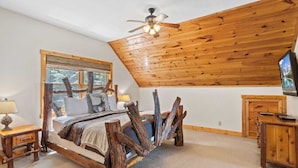 A rustic bedroom with a wooden ceiling, a bed with a deer pillow, a window with a view, and a TV on a wooden stand.