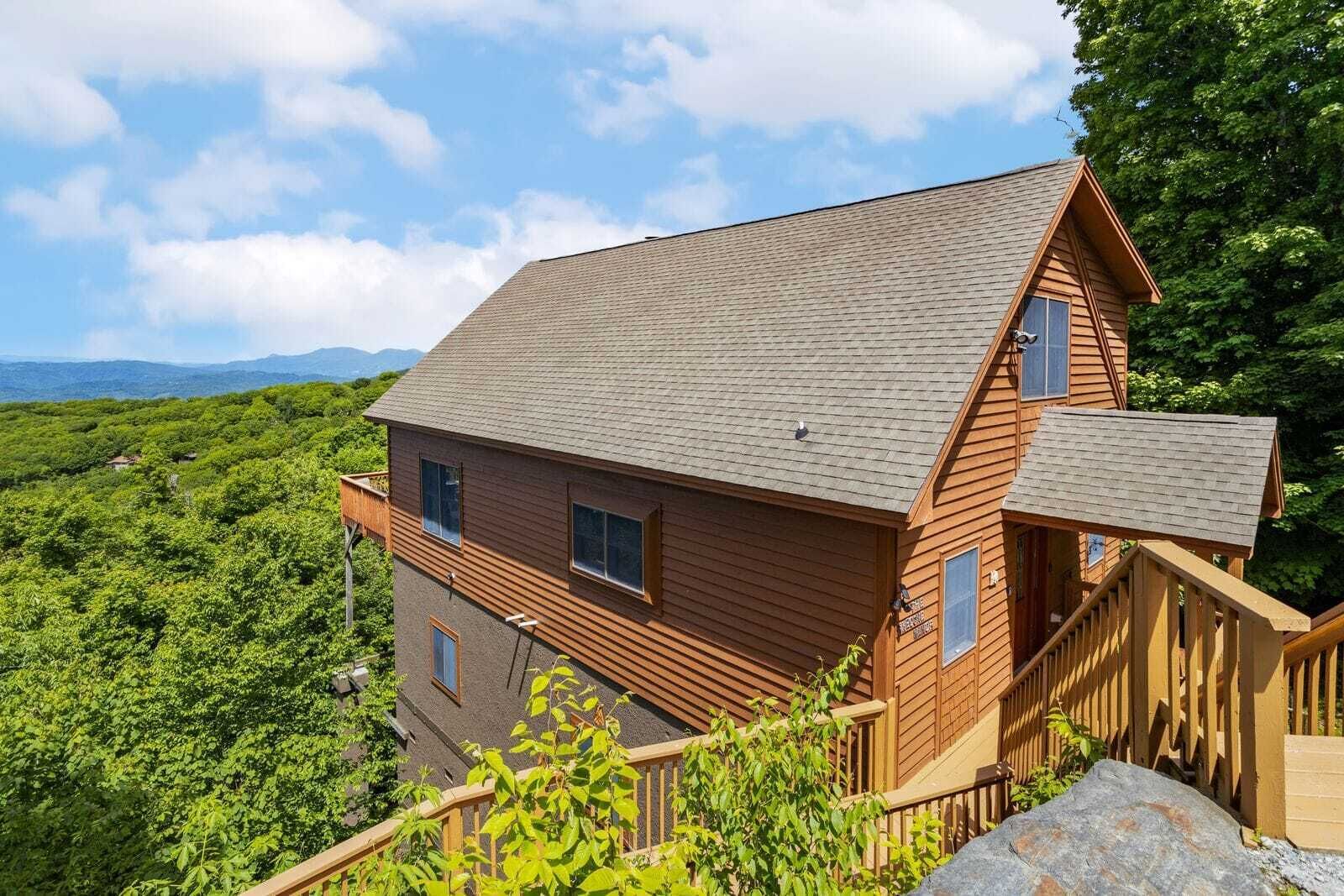 A wooden cabin with a sloped roof, wooden deck, and a view of a lush green forest.