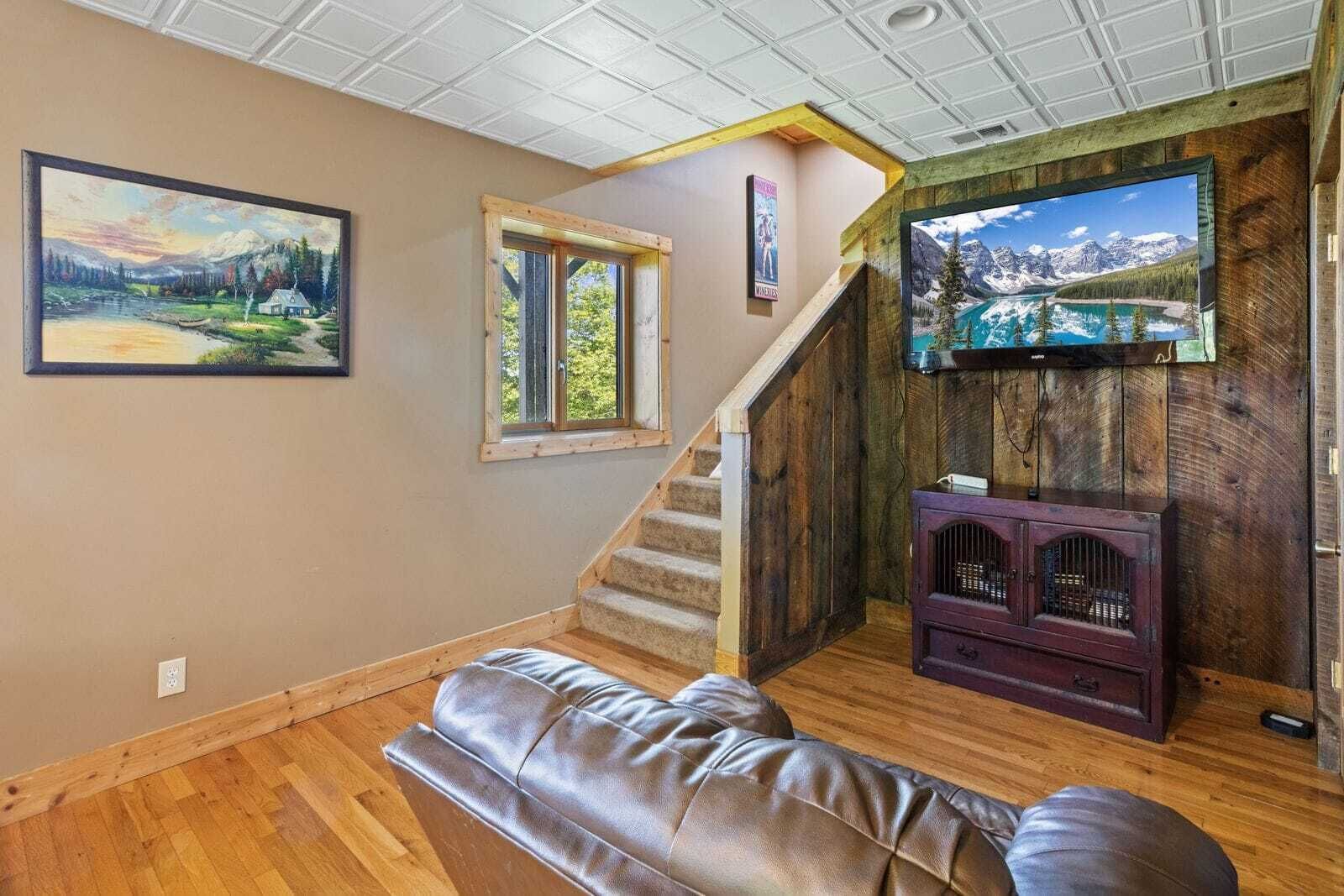 A cozy living room with a wooden staircase, a leather sofa, a TV mounted on a wooden wall, and a painting of a landscape.
