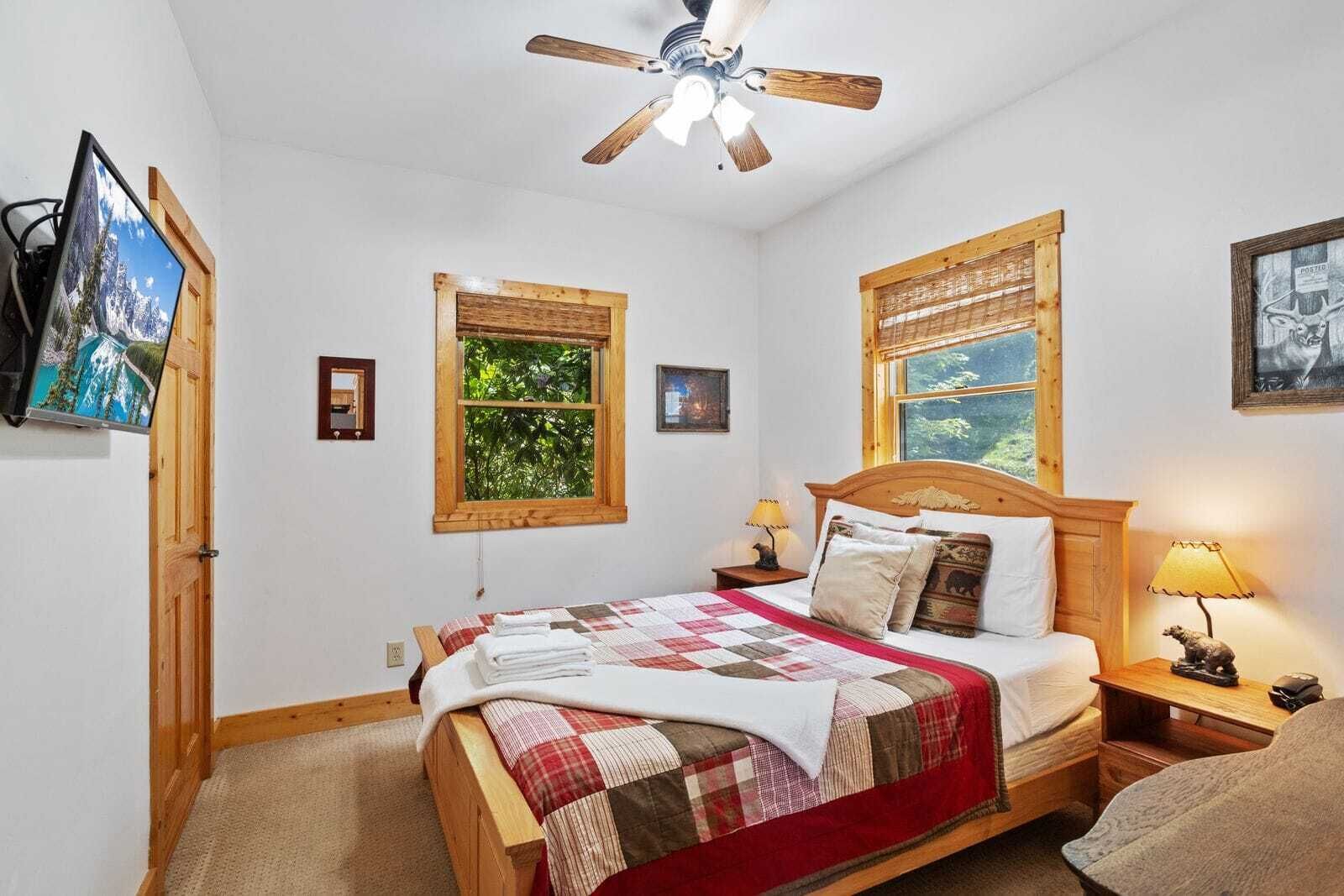 A bedroom with a wooden bed, two windows with wooden frames, a ceiling fan, and a TV mounted on the wall.