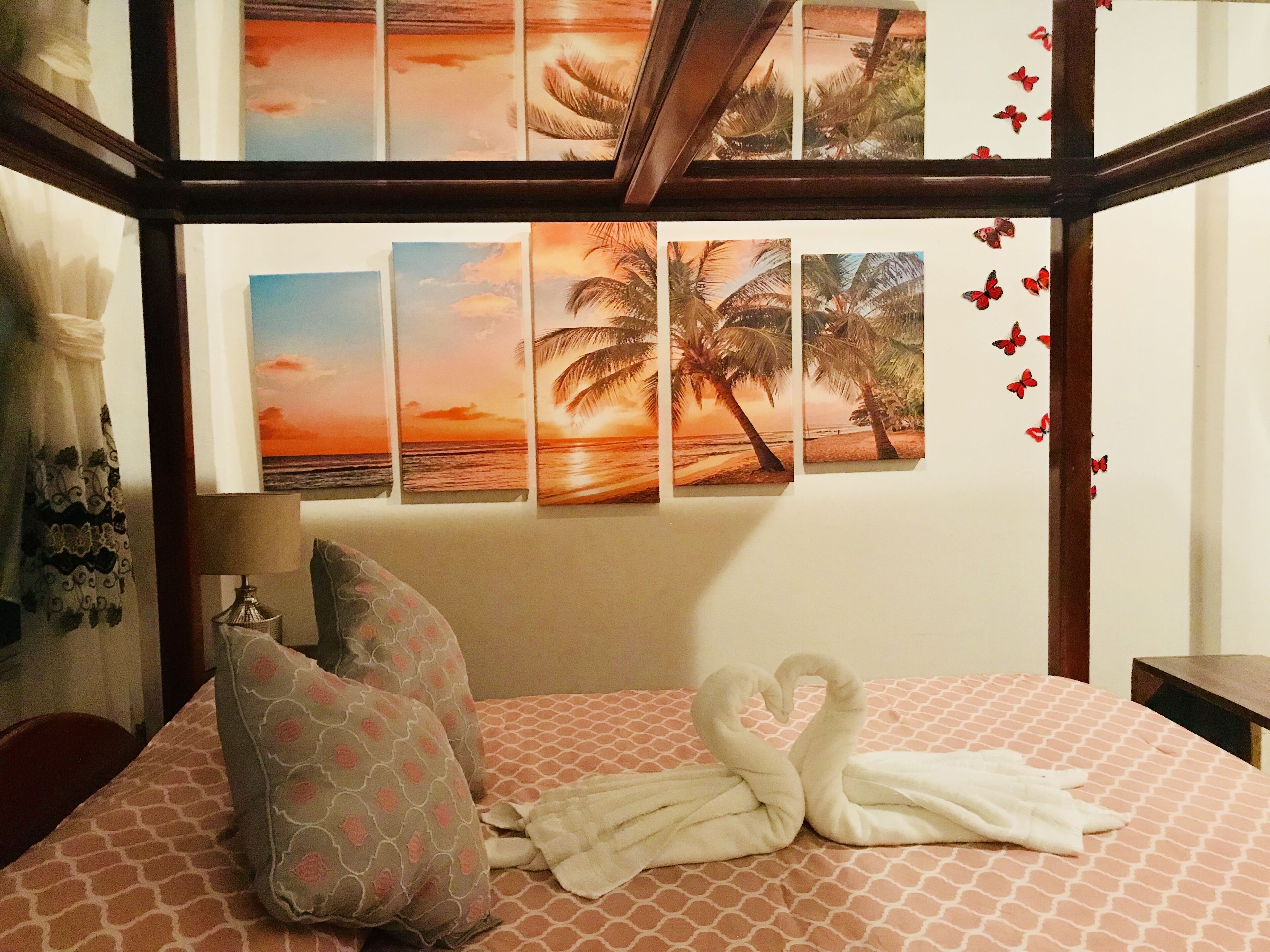 A hotel room with a bed, pillows, a lamp, and a wall with multiple sunset and palm tree paintings.