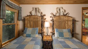 A room with two wooden beds with checkered bedding, a small wooden table with a lamp, and a window with plaid curtains.