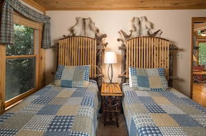 A room with two wooden beds with checkered bedding, a small wooden table with a lamp, and a window with plaid curtains.