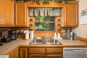 A kitchen with wooden cabinets, a double sink, a coffee maker, and a window with a view of greenery.