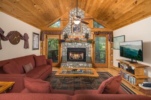 A cozy living room with a stone fireplace, wooden ceiling, and a large television.