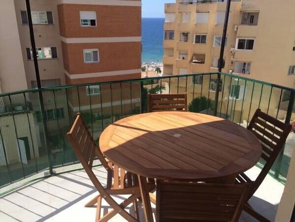 A balcony with a wooden table and chairs, overlooking a beach and other buildings.