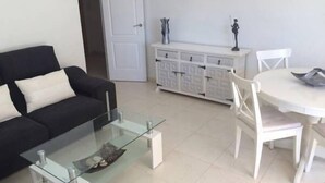 A living room with a black sofa, a glass coffee table, a white dining table with chairs, and a white cabinet with drawers. - Palasiet (Villajoyosa)