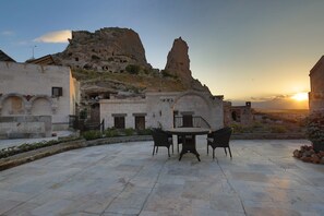 A courtyard with a stone table and chairs, a stone wall, and a view of a rocky hill with a tall rock formation. - Romance Cave Suites (Nevsehir)