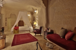 A bedroom with a canopy bed, a red sofa, a red rug, a wooden chair, and a small table with a lamp. - Romance Cave Suites (Nevsehir)