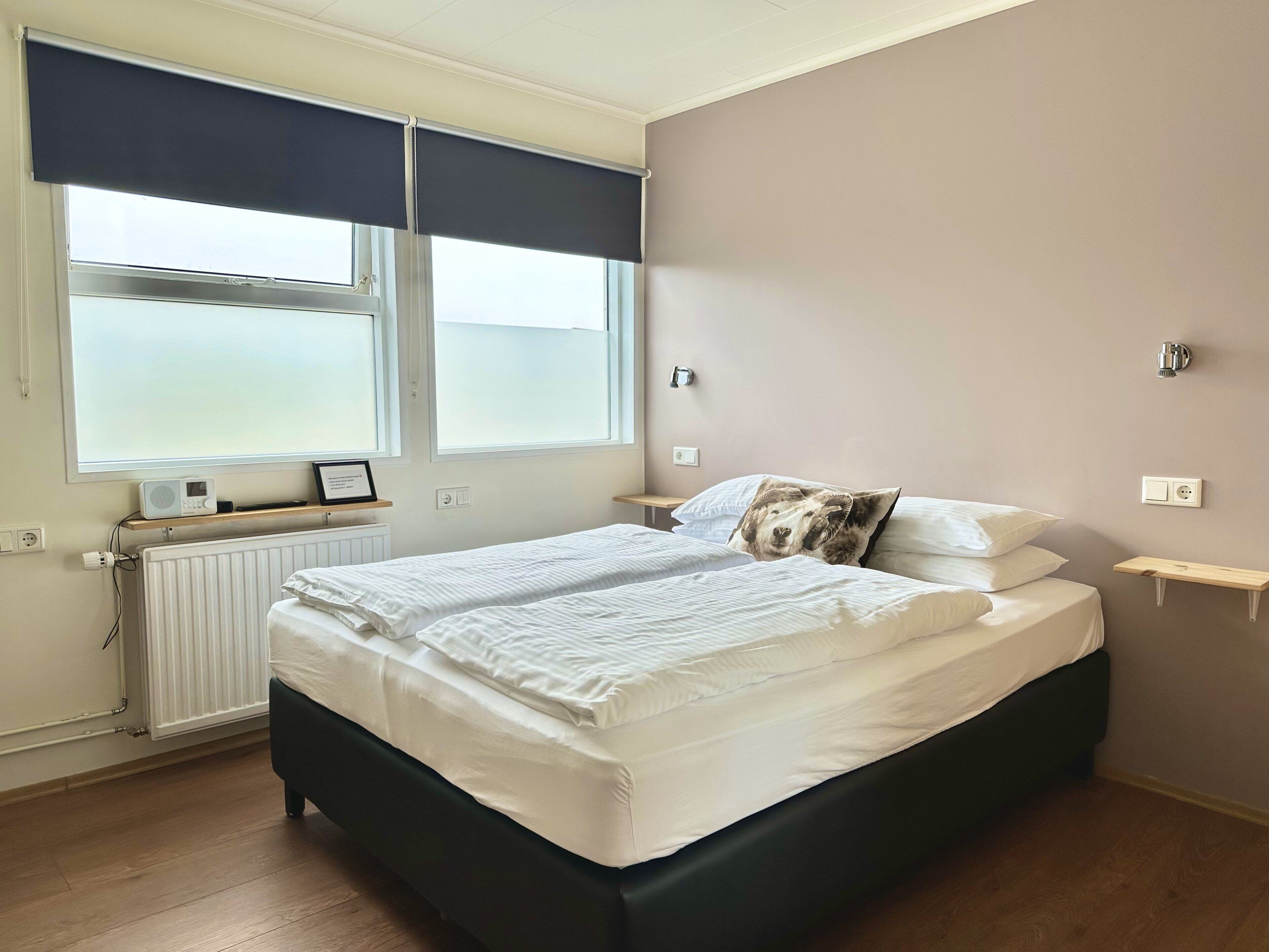 Un dormitorio con cama, mesitas de noche, un radiador y una ventana con persianas.