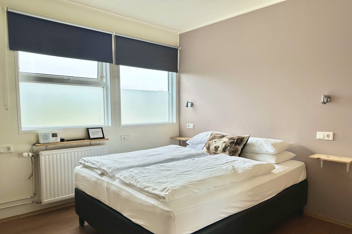 Ein Schlafzimmer mit Bett, Nachttischen, Heizkörper und einem Fenster mit Jalousien.