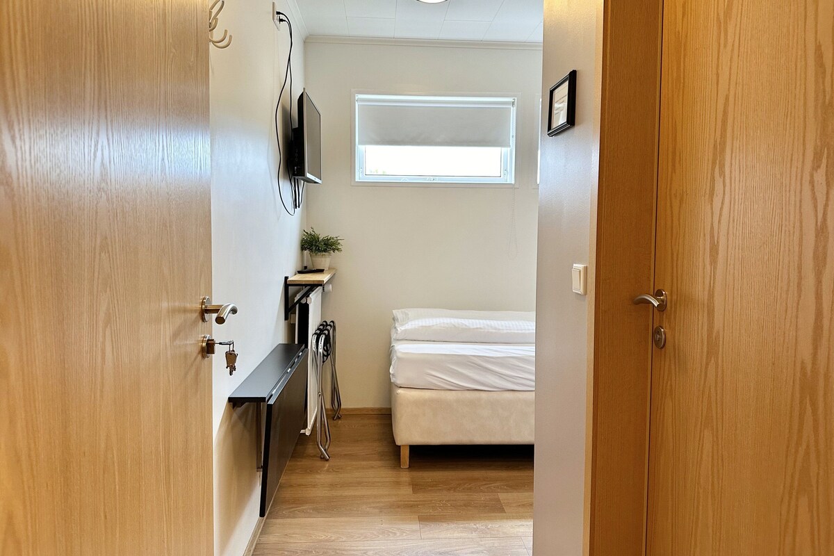 Ein schmaler Flur führt zu einem Schlafzimmer mit einem Bett, einem kleinen Tisch mit einer Pflanze, einem an der Wand montierten Fernseher und einem Fenster, das natürliches Licht hereinlässt.