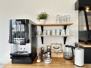 Une machine à café, des étagères avec des tasses et des mugs, une boîte de rangement et une bouilloire sur une table en bois.