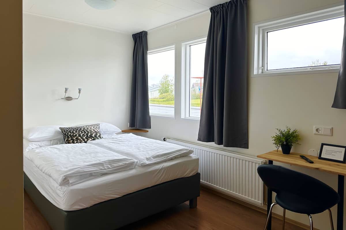 Ein Schlafzimmer mit Bett, Schreibtisch, Stuhl, einem Fenster mit Vorhängen, einem Heizkörper und einer Deckenleuchte.