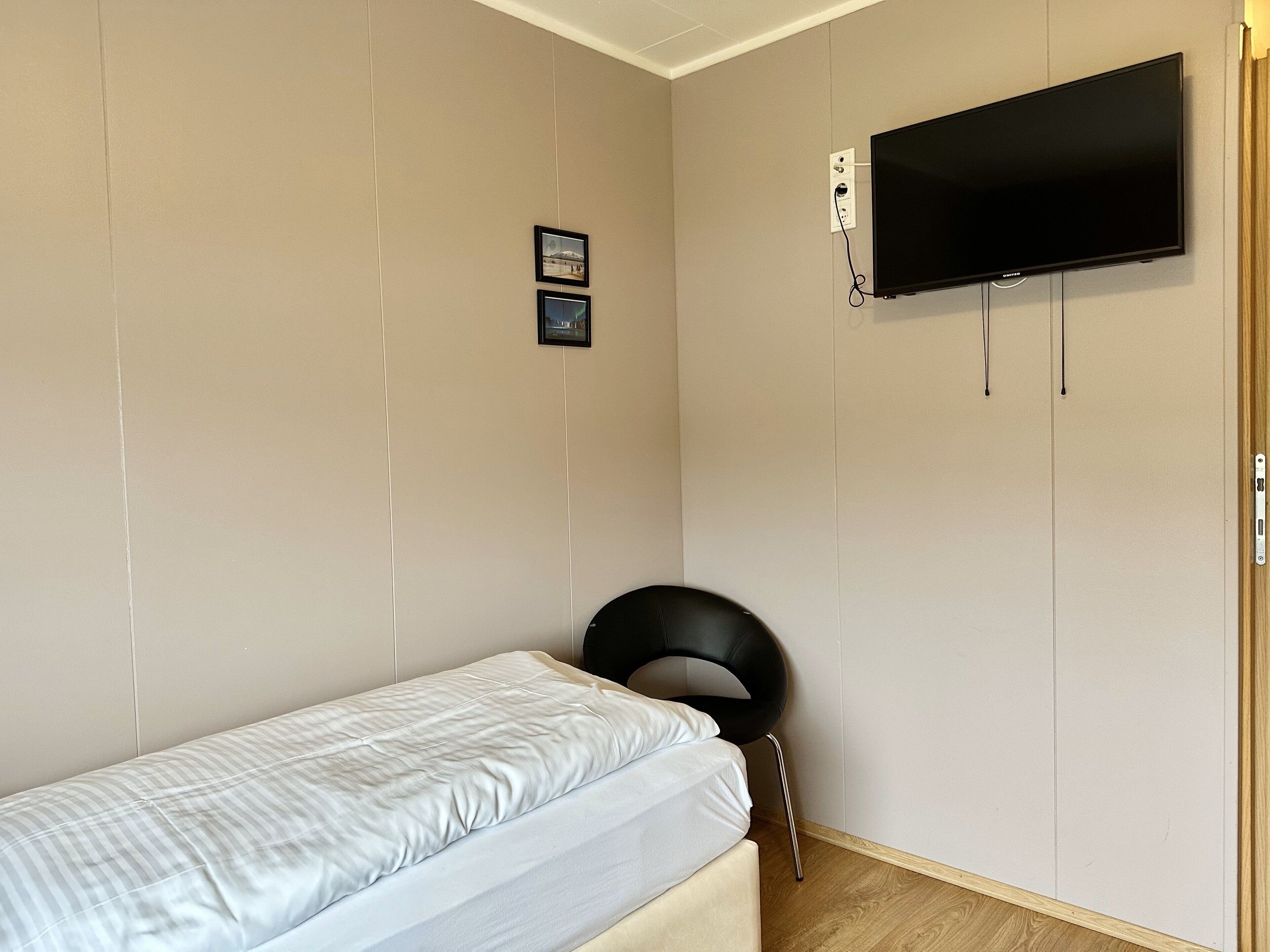 Un dormitorio pequeño y ordenado con una cama individual, un televisor montado y una silla.