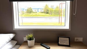 Ein Hotelzimmer mit Blick aus dem Fenster auf ein Gebäude und Bäume, einer Zimmerpflanze, einem gerahmten Willkommensschild und einer Fernbedienung auf dem Nachttisch.