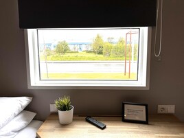 A hotel room with a window view of a building and trees, a bedside plant, a framed welcome sign, and a remote control on the nightstand.